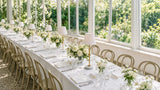 Marble-base table lamps with white shades and flowers along a wedding table in a sunlit conservatory