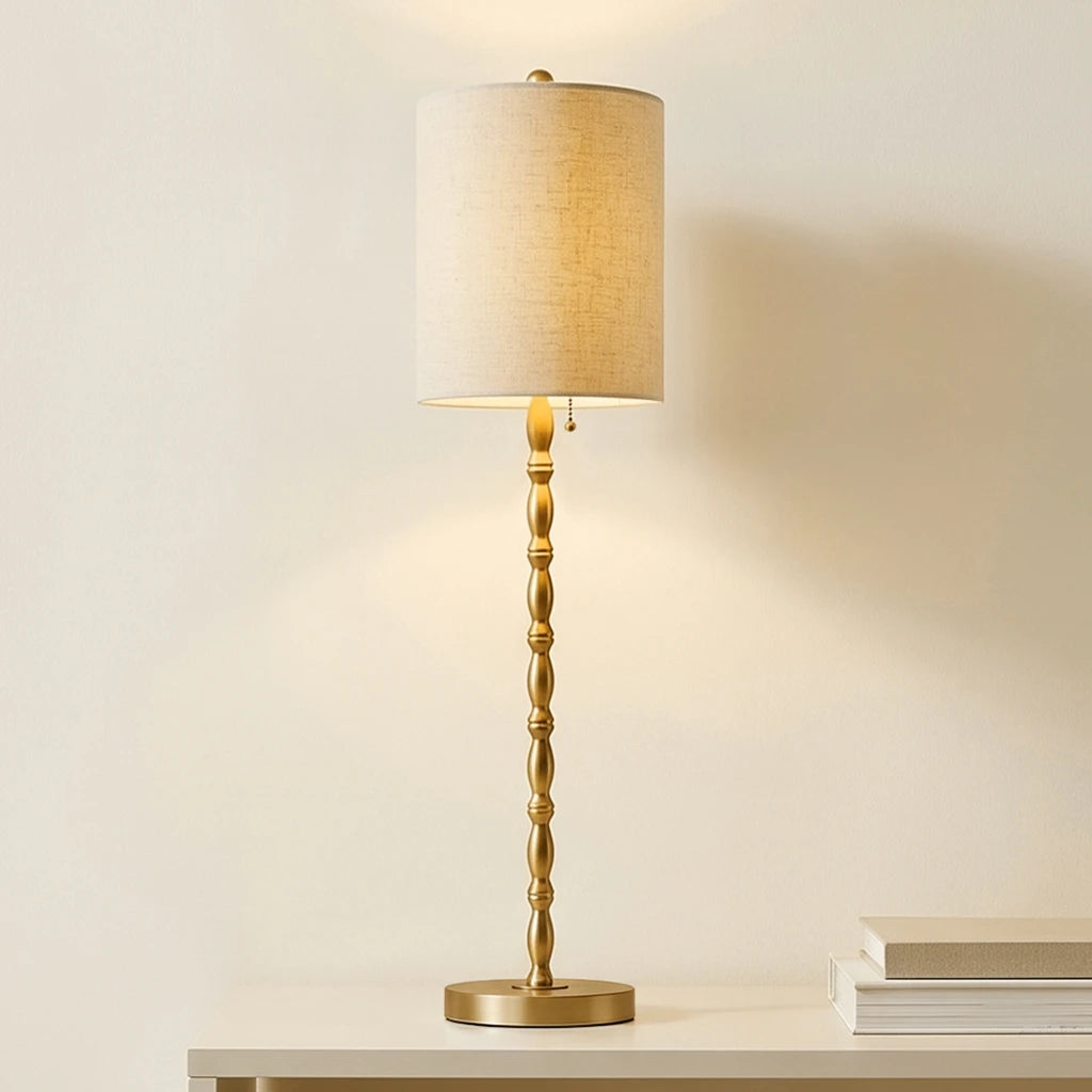 Single Aurelia brass-finish buffet lamp with a linen drum shade glowing on a console beside stacked books.