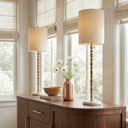 Pair of Noor brass-finish buffet lamps with white marble bases on a wood sideboard by sunlit windows.