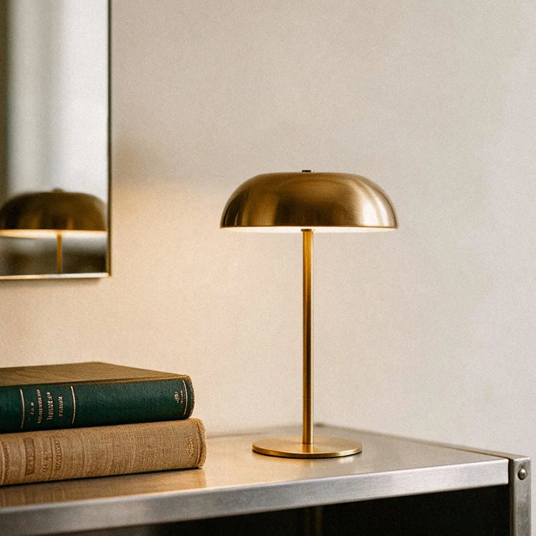Brushed brass cordless mushroom table lamp glowing beside vintage books and a wall mirror.