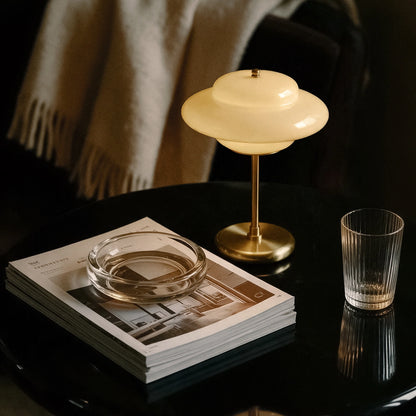 Lampworks glass table lamp glowing on a black side table beside stacked magazines and a drinking glass